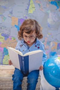 girl reading book while sitting beside desk globe