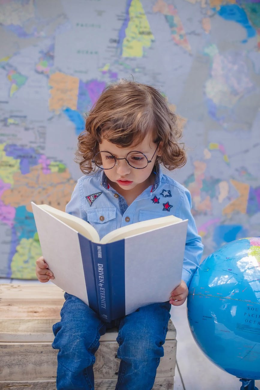 girl reading book while sitting beside desk globe